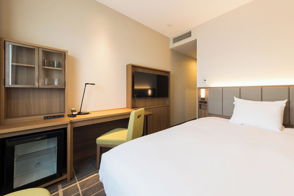 A hotel room with a bed, desk, chair, television, and a small kitchenette.