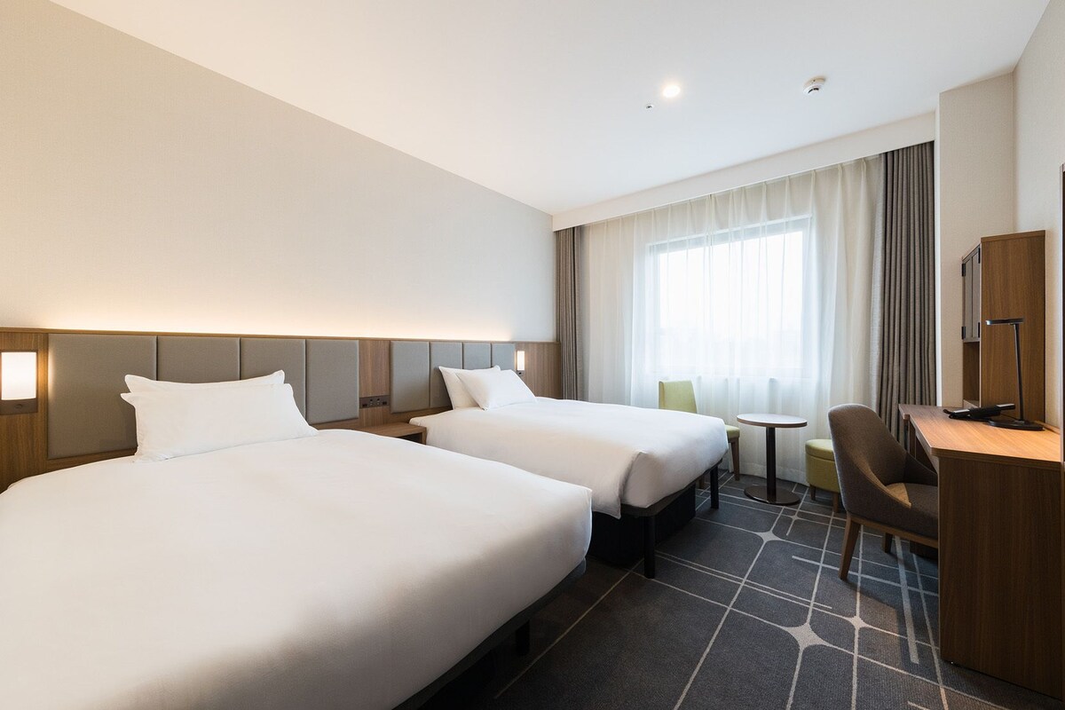 A hotel room with two beds, a desk, a chair, a television, and a window with curtains.