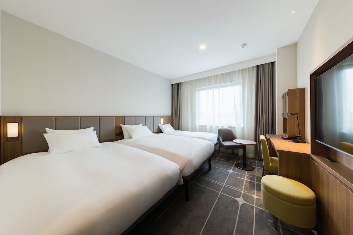 A hotel room with two large beds, a desk, a chair, a TV, and a window with curtains.