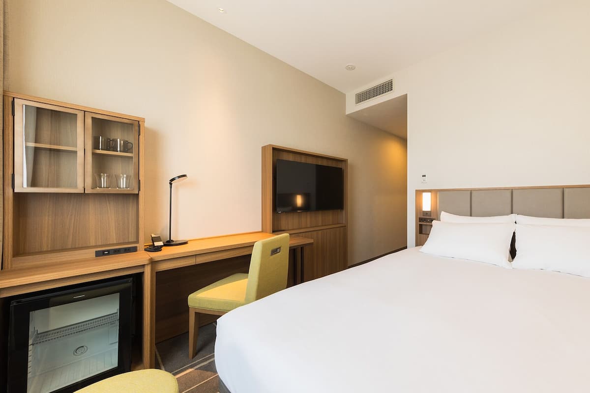 A hotel room with a bed, desk, chair, television, and a minibar.