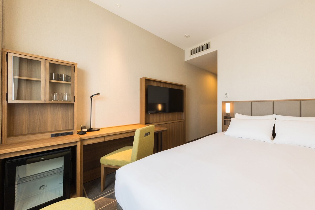 A hotel room with a bed, desk, chair, television, and a minibar.