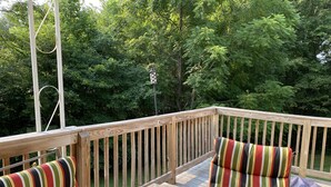 Eine Holzterrasse mit Metallgeländer, zwei gestreifte Sitzstühle, ein Metallkübel und Blick auf dichtes Grünwerk.