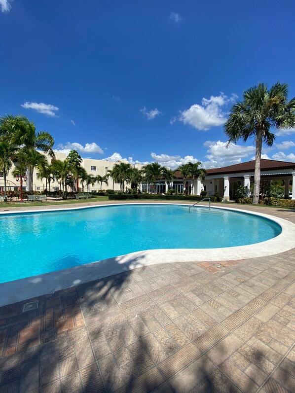A swimming pool with a tiled deck, surrounded by palm trees and buildings with a clear blue sky above.