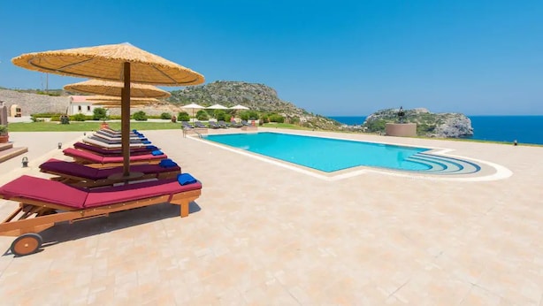 Ein Poolbereich mit Liegestühlen und Sonnenschirmen, umgeben von einem angelegten Garten und Blick auf das Meer.