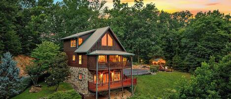 A two-story log cabin with a balcony, surrounded by dense forest, during sunset.
