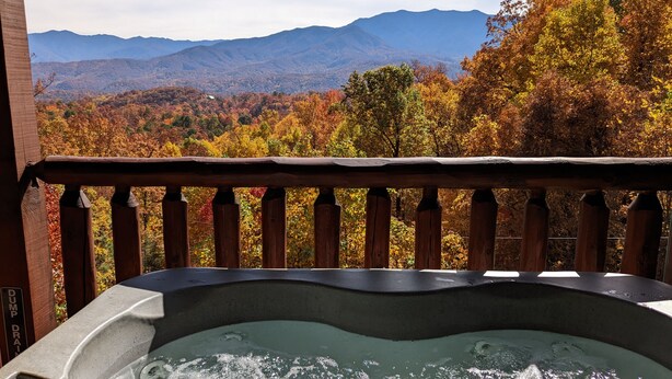 A hot tub with a wooden deck and a view of autumn foliage and mountains.