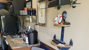 Une cuisine compacte équipée d’un four à micro-ondes, d’une cuisinière et d’un évier. On y trouve une petite table avec une chaise et un panier rempli de livres. Un téléviseur est fixé au mur, au-dessus du comptoir.