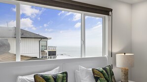 A bedroom with a view of the sea, a white bed with two pillows, a nightstand with a lamp, and a window with a view of a building and the ocean.