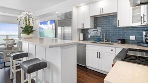 A modern kitchen with white cabinets, a stainless steel refrigerator, a dishwasher, and a large island with bar stools.