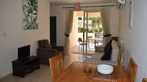 Een woonkamer met een televisie, bank en een eettafel met stoelen. Er is een balkon met uitzicht op palmbomen.