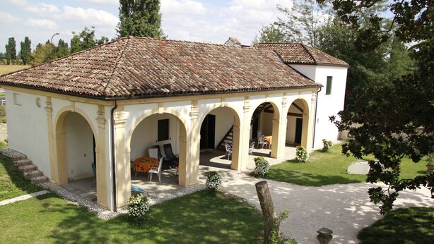A small, single-story building with a tiled roof and arched doorways, surrounded by a well-maintained lawn and a gravel path. - Magnolia, charming residence in the Barchessa of Venetian Villa: Villa Pastori (Mira)