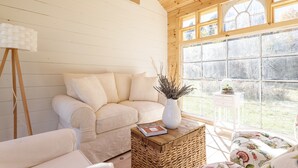 Una stanza accogliente con soffitto in legno, un divano beige, una poltrona bianca, un tavolino con un libro e un'ampia finestra con vista sugli alberi.