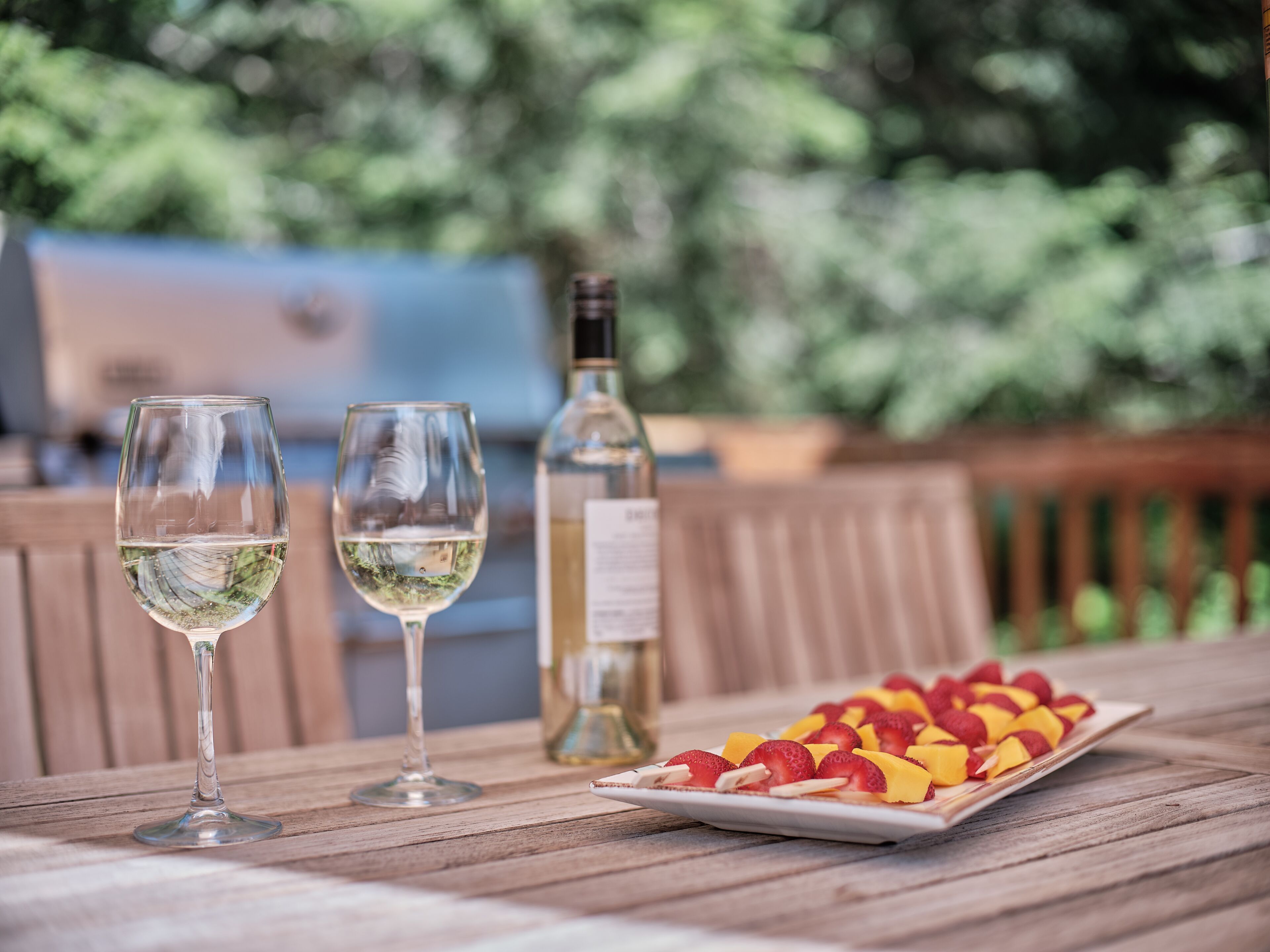 Zwei Weingläser auf einem Holztisch mit einer Flasche Weißwein und einer Schale Obstscheiben.