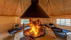 Interno di una baita in legno con una stufa a legna al centro, una panca con un cuscino a forma d'orso e una finestra vista all'esterno.
