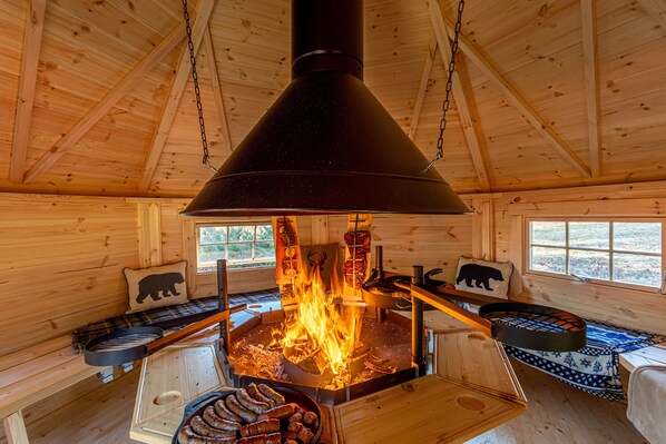 Interior de una cabaña de madera con un fogón central, un banco con un cojín de oso y una ventana con vistas al exterior.