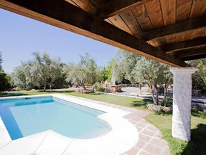 A swimming pool with a wooden pergola overhead, surrounded by a stone pathway and greenery.
