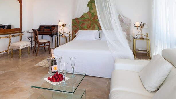Ein Schlafzimmer mit Bett, einem Glastisch mit Flasche und Gläsern, einem Sessel, einem Schreibtisch und einem Spiegel.