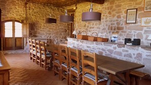 Un coin repas rustique aux murs de pierre, avec des bancs en bois et une longue table, agrémenté de lampes suspendues et d’une étagère où sont posés des paniers et des livres.