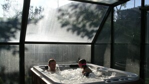 Un bain à remous où se trouvent deux personnes, entouré d’une enceinte de verre offrant une vue sur les arbres et une route à l’extérieur.