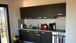 Een moderne keuken met donkere kasten, een aanrecht en een fornuis. Er staat een prullenbak en een raam met zonwering.
