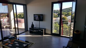 Een moderne woonkamer met een flatscreen televisie op een standaard, een klein tafeltje met een vaas, en uitzicht op een balkon met een hekwerk.