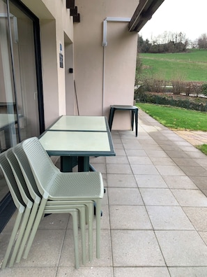 A patio area with a table and chairs, a small stool, and a view of a green field. - Cottage 5 people on the quiet riverside le Lizon
in Franche Comté (Nans-Sous-Sainte-Anne)