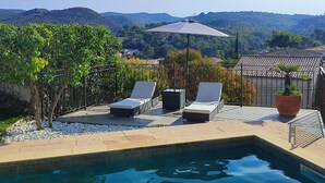 Piscina con sillas de descanso y una planta en maceta, rodeada por una valla y árboles, con vistas a colinas al fondo.