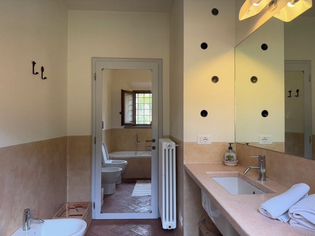 Un baño con un lavabo blanco, un espejo y un radiador. Hay una puerta que da a otra habitación con un inodoro y una ventana.