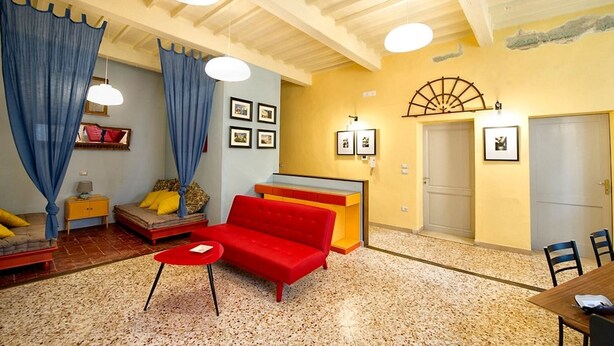 A cozy living room with a red sofa, a small red table, and a yellow cabinet. There are blue curtains, a wooden ceiling, and a wall with framed pictures.