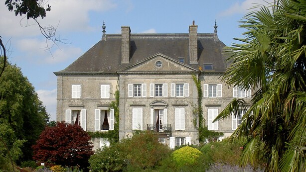 Un grand bâtiment historique à la façade symétrique, doté de nombreuses fenêtres avec volets et d’une entrée centrale.