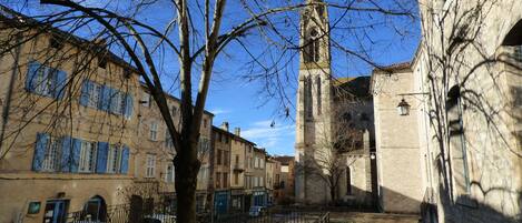 A courtyard with a church tower, buildings with blue shutters, and a tree in the foreground.