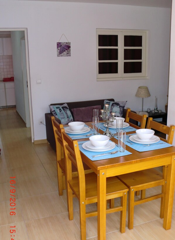 A dining area with a wooden table set for four, surrounded by chairs. A sofa is positioned against the wall, and a window with a white frame is visible above the table.