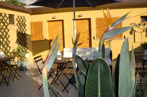 An outdoor patio with a wooden table and chairs, surrounded by large green plants and a yellow wall with wooden doors.