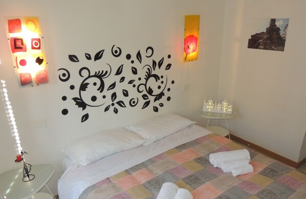 A neatly made bed with white linens, a colorful checkered bedspread, and two rolled towels on the floor. There is a small table with a lamp and a decorative item on it, and a wall-mounted light fixture with a floral design. - Esquiliae apartment (Roma)