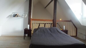 Een slaapkamer met een schuine kap, houten balken, een bed met donkere dekbedovertrek, een nachtkastje met een lamp en een kledingrek.