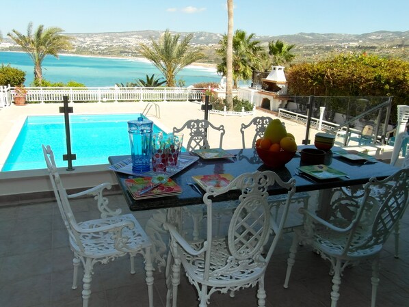 A dining table with chairs, a fruit bowl, and a view of the sea and hills.