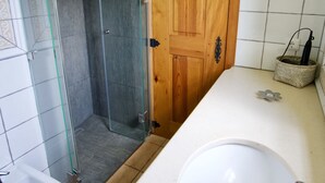 Um banheiro com box de vidro, porta de madeira, pia branca e uma pequena cesta no balcão.