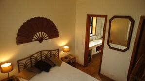 Quarto de hotel com cama, luminárias de cabeceira, ventoinha na parede, espelho e banheiro visível por uma porta aberta.