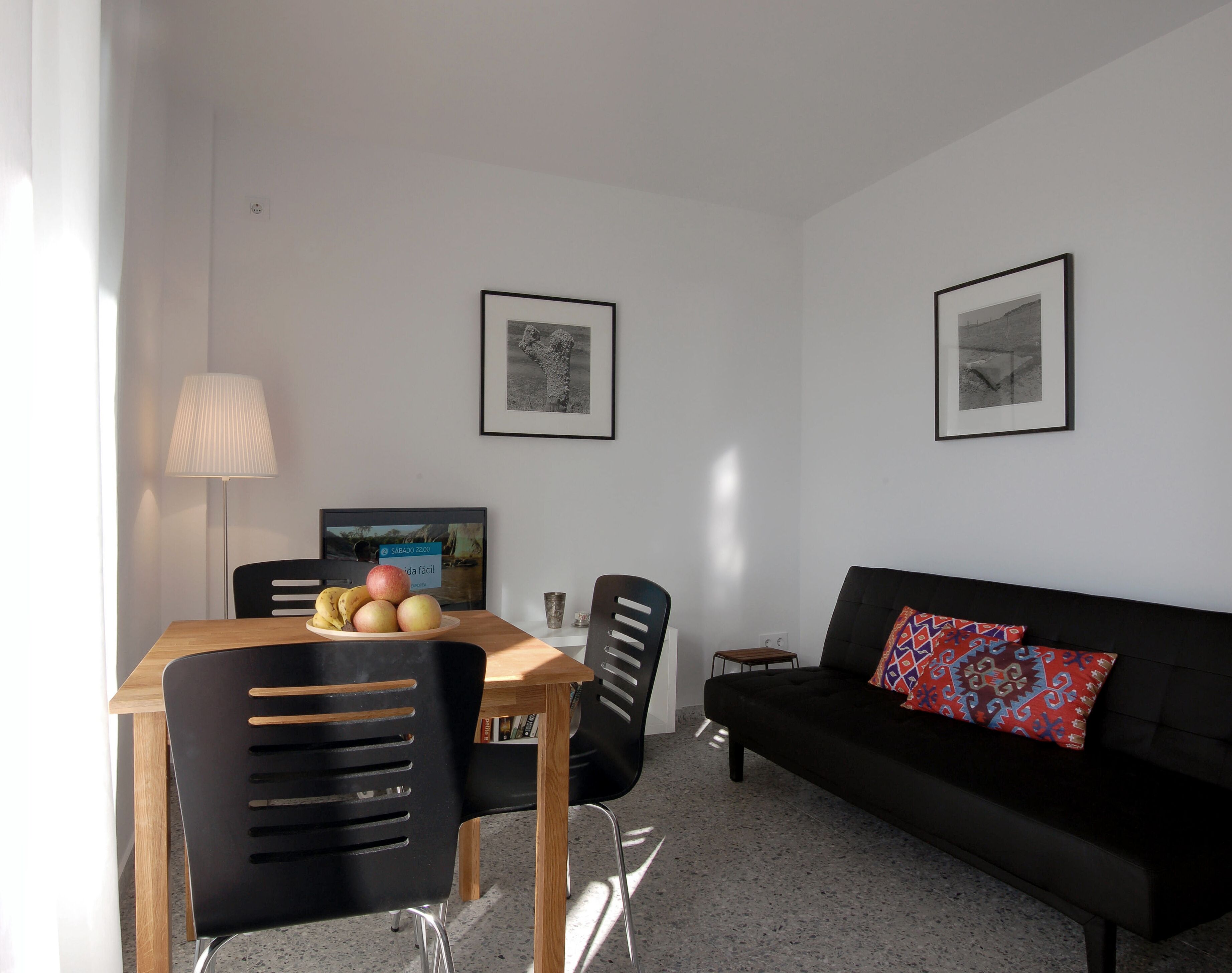 A modern living room with a wooden dining table, black chairs, a black sofa with patterned cushions, and two framed pictures on the wall.