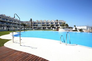 A swimming pool with a wooden deck, a white building with balconies in the background, and a clear blue sky. - Blue Fin, your unfogettable getaway in Tarifa! ☀️ (Tarifa)