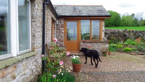 A stone house with a wooden door and windows, a paved garden area with flowers, and a black dog standing outside. - Pet friendly barn conversion with spectacular views, just for 2. (Crosby Garrett)