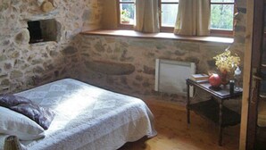 Une pièce aux murs de pierre, avec un lit, une fenêtre avec des rideaux, un radiateur et une petite table sur laquelle se trouvent un vase et un livre.