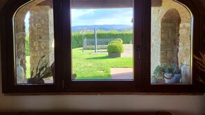 A view through a wooden-framed window with stone walls, revealing a well-maintained garden with a metal fence, potted plants, and a clear sky. - Stone house with private exclusive infinity pool in Chianti (San Filippo A Ponzano)