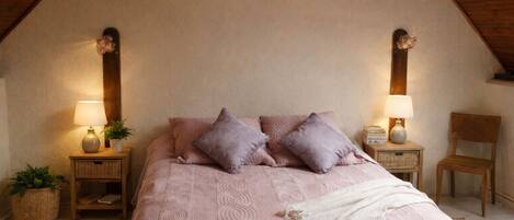 Une chambre avec un plafond en pente, un lit avec une literie rose, deux tables de chevet avec des lampes, une chaise et une petite plante.