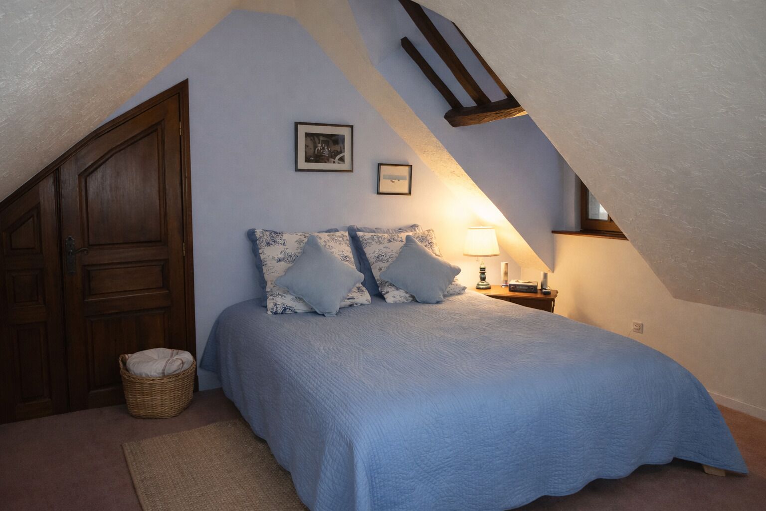 A bedroom with a sloped ceiling, a bed with blue bedding, two pillows, a bedside lamp, a framed picture, and a wooden wardrobe.