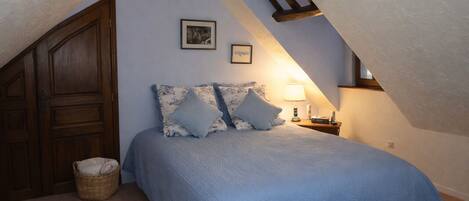 A bedroom with a sloped ceiling, a bed with blue bedding, two pillows, a bedside lamp, a framed picture, and a wooden wardrobe.