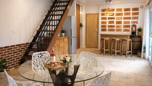 Ein Essbereich mit Glastisch und weißen Stühlen, eine Holztreppe, eine Küche mit Holzarbeitsplatte und eine Wand mit Holztafeln.
