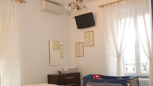 Un dormitorio con cama, un escritorio con computadora, un televisor montado en la pared y una ventana con cortinas.