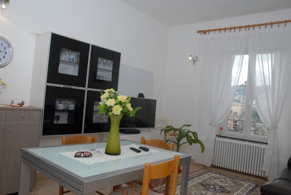A dining area with a table, chairs, a vase of flowers, a clock, a television, and a window with curtains.