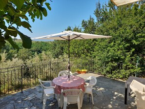 Un patio al aire libre con una mesa, sillas, un paraguas blanco y vistas a una zona boscosa.